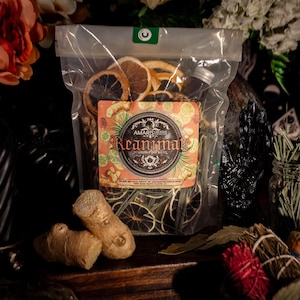 May include: A clear plastic bag filled with dried orange and lime slices, herbs, and a small bottle. The bag has a label with the word "Reanimate" on it. Ginger root and bundles of herbs are in front of the bag. A jar of herbs and an orange are in the background.