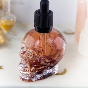 May include: A clear, skull-shaped glass bottle with a black dropper top. The bottle is filled with a reddish-brown liquid. Other bottles and a golden skull are in the background. The bottle is on a white surface.