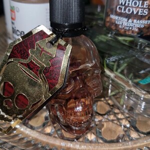 May include: A skull-shaped glass bottle with a black dropper top and a gold and red tag. The bottle is filled with a brown liquid and sits on a clear, decorative glass plate. Whole cloves are visible in the background.