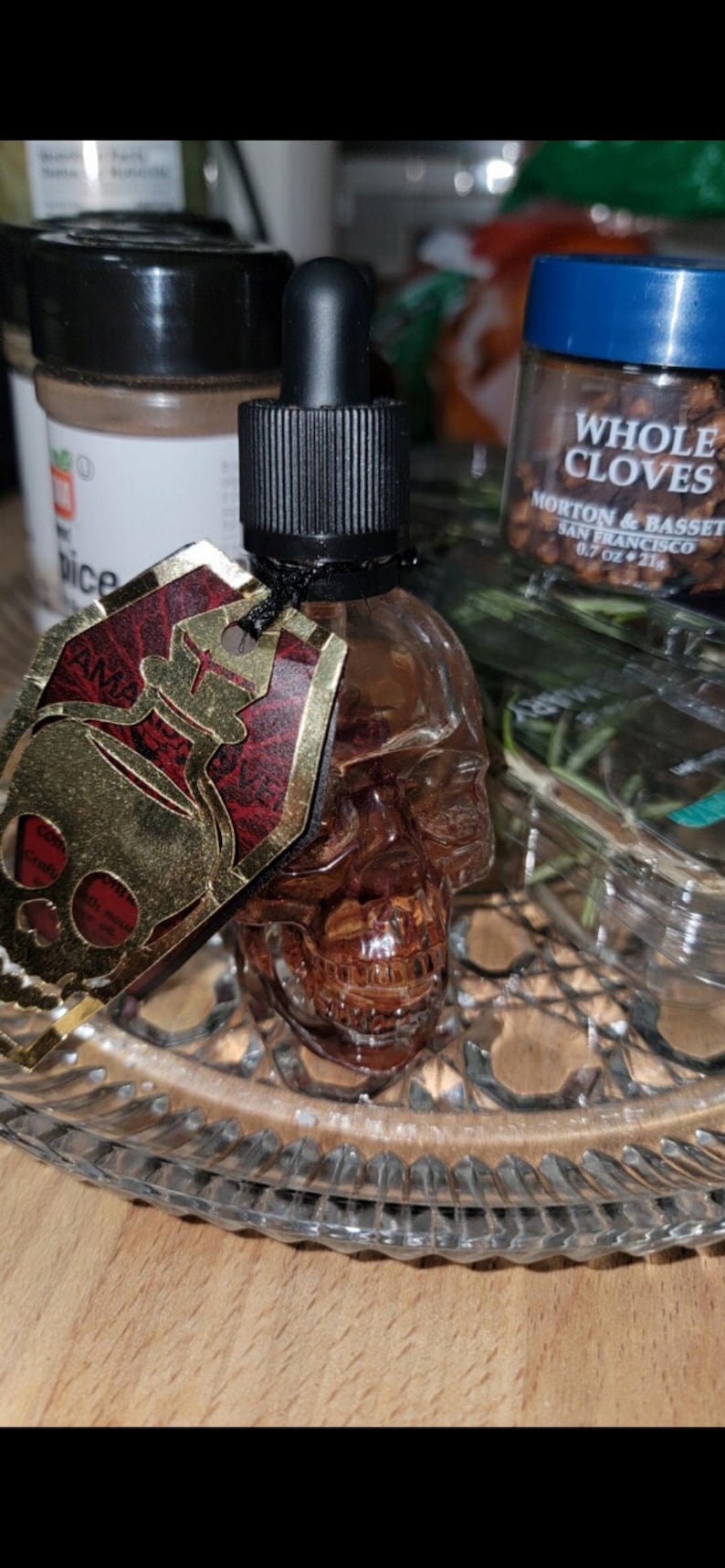 May include: A skull-shaped glass bottle with a black dropper top and a gold and red tag. The bottle is filled with a brown liquid and sits on a clear, decorative glass plate. Whole cloves are visible in the background.