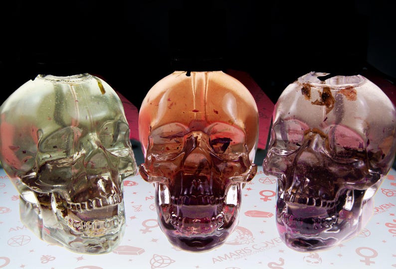 May include: Three transparent skull-shaped decorative objects with varying shades of color. The center skull is a deep reddish-brown, flanked by a green and a purple skull. The base has the text "AMARIS COVE".