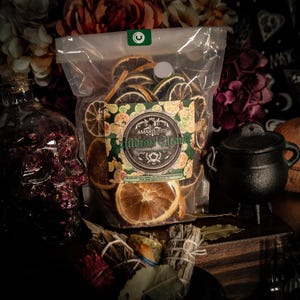 Puede incluir: Una bolsa de plástico transparente llena de rodajas de naranja y lima secas, etiquetada como "Cauldron Cleans". También se ven un pequeño caldero negro, una botella con forma de calavera y manojos de hierbas. El fondo incluye flores.