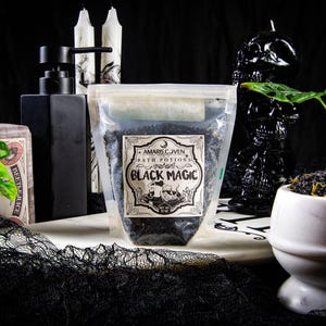 Może przedstawiać: Mroczna martwa natura z torebką z miksturami do kąpieli "Black Magic", czarnym dozownikiem mydła i świecami w kształcie czaszki. Biała marmurowa miska zawiera sole do kąpieli i zieleń. Wszystko na czarnym tle.