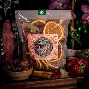 Puede incluir: Una bolsa transparente llena de rodajas de naranja secas, hojas de laurel y palitos de canela. Es visible una etiqueta rosa con la palabra "Smolder". Otros elementos incluyen un cuenco con capullos de rosa secos, una vela roja y una naranja.