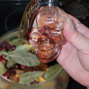 May include: A clear, skull-shaped glass bottle with a black dropper top. A decorative gold and red embellishment is attached to the bottle. The bottle is held in front of a glass bowl filled with botanicals.