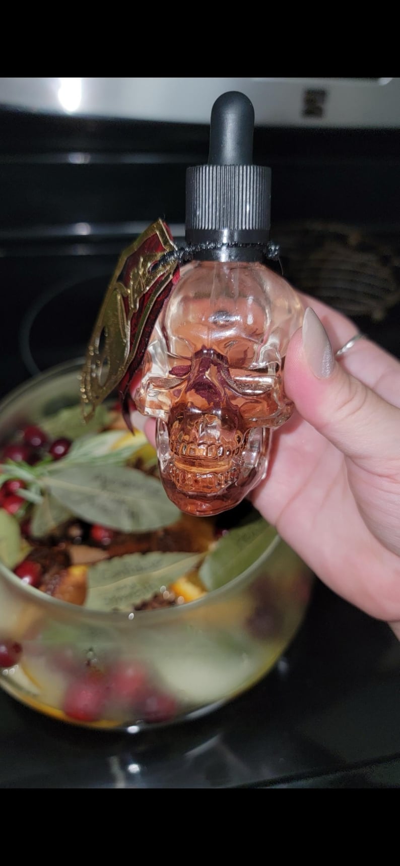 May include: A clear, skull-shaped glass bottle with a black dropper top. A decorative gold and red embellishment is attached to the bottle. The bottle is held in front of a glass bowl filled with botanicals.