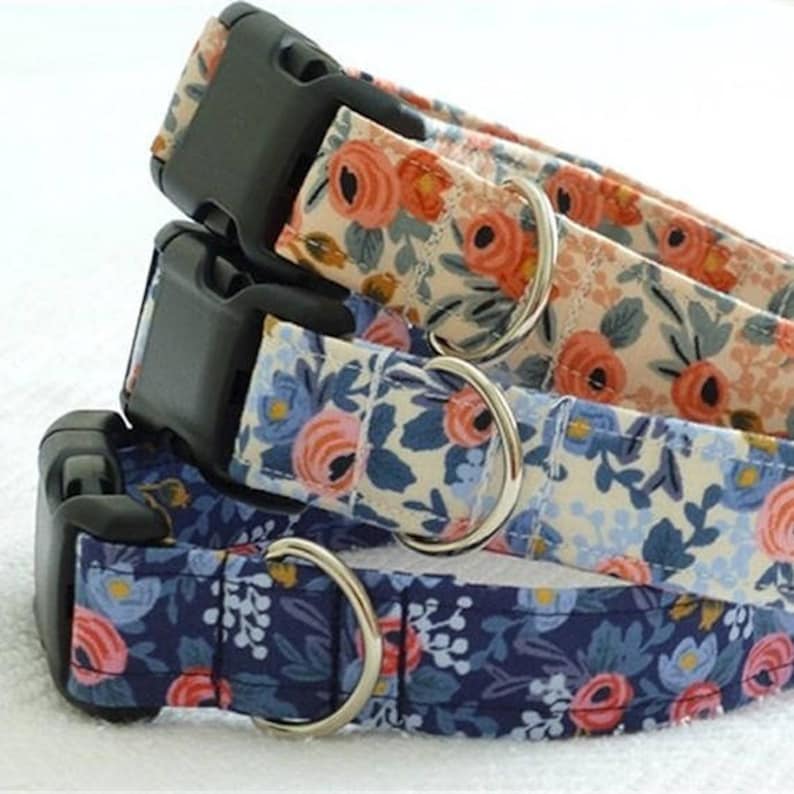 May include: Three fabric collars stacked vertically, each featuring a floral pattern with roses and leaves. The collars have black plastic side-release buckles and silver-toned metal D-rings. The top collar is cream, the middle is light blue, and the bottom is navy blue.