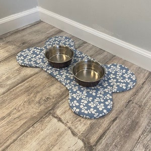 May include: A bone-shaped fabric mat featuring a blue background with a white leaf pattern. Two circular stainless steel bowls are placed on top of the mat. The mat is designed to hold pet food or water dishes, providing a stable surface for feeding.