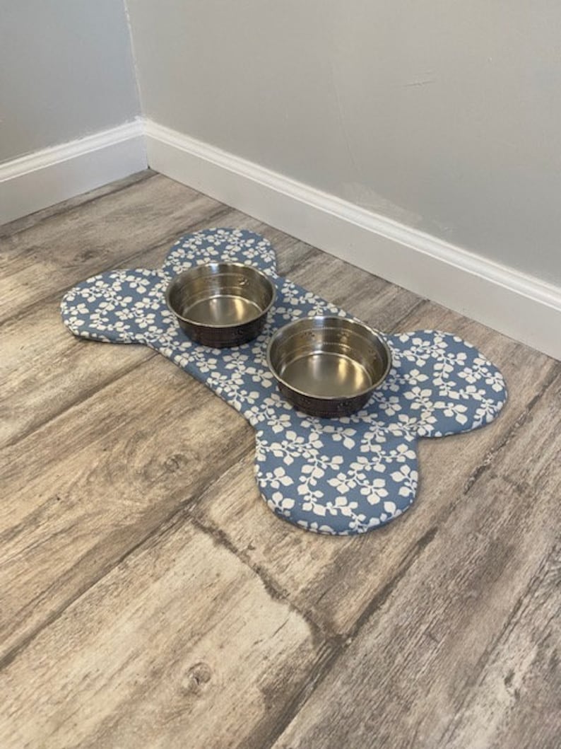 May include: A bone-shaped fabric mat featuring a blue background with a white leaf pattern. Two circular stainless steel bowls are placed on top of the mat. The mat is designed to hold pet food or water dishes, providing a stable surface for feeding.