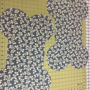 May include: Two fabric cutouts shaped like dog bones, featuring a blue background with a repeating white leaf and vine pattern. The pieces are laid flat on a green cutting mat with black grid lines and inch markings along the bottom edge.