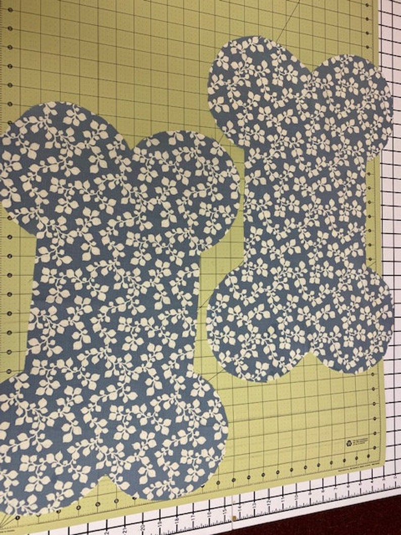 May include: Two fabric cutouts shaped like dog bones, featuring a blue background with a repeating white leaf and vine pattern. The pieces are laid flat on a green cutting mat with black grid lines and inch markings along the bottom edge.