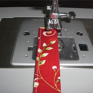 May include: A red fabric strip with a white and green floral pattern is positioned under the silver presser foot of a sewing machine. The fabric features a straight white stitched seam running along the edge, while the metal needle plate shows visible measurement markings in inches.