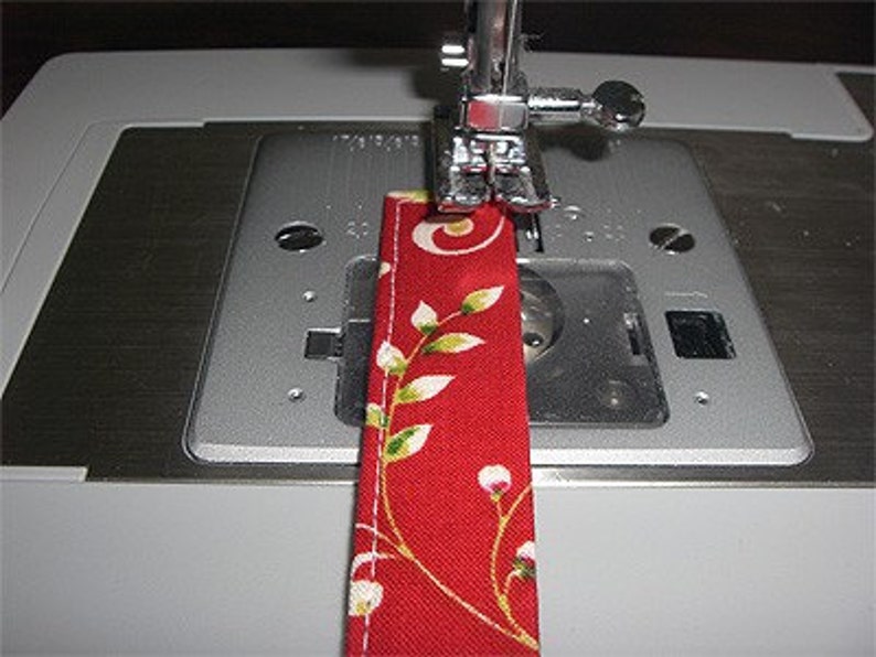 May include: A red fabric strip with a white and green floral pattern is positioned under the silver presser foot of a sewing machine. The fabric features a straight white stitched seam running along the edge, while the metal needle plate shows visible measurement markings in inches.
