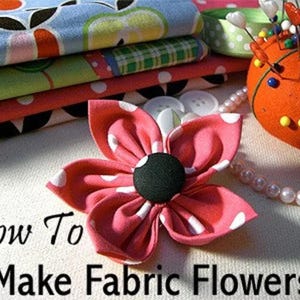 Puede incluir: Una flor de tela con p&eacute;talos de lunares rosas y blancos y un bot&oacute;n central negro s&oacute;lido. El texto "How To Make Fabric Flowers" est&aacute; impreso debajo de la flor. Se ven pilas de tela estampada, un alfiletero verde y cuentas blancas.