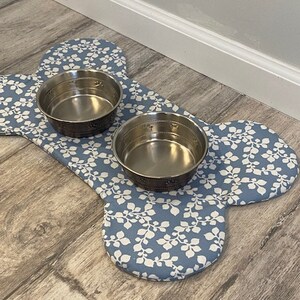 May include: A bone-shaped pet mat featuring a blue fabric with a white leaf pattern, holding two circular stainless steel bowls. The mat is designed with a soft, padded surface and provides a stable base for the two metallic feeding dishes.