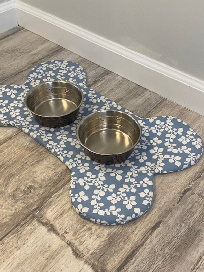 May include: A bone-shaped pet mat featuring a blue fabric with a white leaf pattern, holding two circular stainless steel bowls. The mat is designed with a soft, padded surface and provides a stable base for the two metallic feeding dishes.