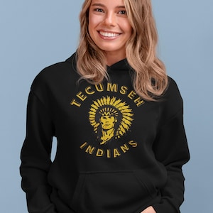 May include: Black hoodie with a gold graphic of a Native American chief and the words "TECUMSEH INDIANS". The hoodie has a front pocket and a drawstring hood. The person is smiling.