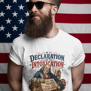 Puede incluir: Camiseta blanca con el texto "Declaration of Intoxication" y una ilustración de Benjamin Franklin con una cerveza. El fondo es una bandera americana.