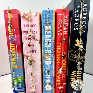 Puede incluir: Cinco marcapáginas de cadena metálica vertical con varios dijes, incluyendo un sombrero de vaquero, botas, una rosa, un dragón y símbolos celestiales, cada uno unido a la parte superior de diferentes lomos de libros como Just for Summer e Iron Flame.