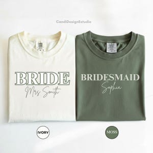 May include: Two t-shirts, one ivory and one moss green, are displayed. The ivory shirt reads "BRIDE Mrs. Smith" in green lettering. The moss green shirt reads "BRIDESMAID Sophia" in white lettering. The shirts are folded and presented on a white background.