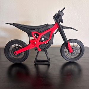 以下が含まれることがあります： 赤と黒のミニチュアバイクモデル。赤いフレーム、黒いタイヤ、シート、ハンドルバーを備えています。バイクは黒いスタンドに展示されており、そのデザインと詳細が強調されています。このモデルは、洗練されたモダンな美学を持っています。