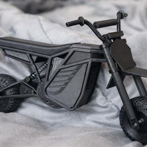 Könnte beinhalten: Ein mattschwarzes Miniaturmotorradmodell mit detailliertem Design. Das Spielzeugmotorrad verfügt über einen Sitz, einen Lenker und robuste Reifen. Das Modell befindet sich vor einem weichen, grauen Hintergrund.