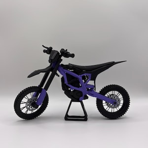 May include: A miniature dirt bike model featuring a vibrant purple frame and black components, including the seat, handlebars, and knobby tyres. The motorcycle is supported by a black stand and displays detailed brake discs on both wheels.
