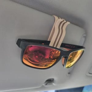 May include: Black sunglasses with orange and yellow lenses are clipped to a car visor with a beige and brown striped clip. The sunglasses are in focus, with the visor and car interior in the background.