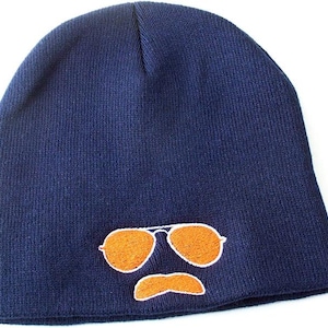 May include: A navy blue beanie hat with an embroidered design of orange aviator sunglasses and a matching mustache. The hat is made of a soft, knitted material, perfect for keeping warm.