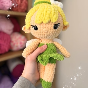May include: A handmade crochet doll with yellow hair, green dress, and small white wings. The doll has black eyes and is held in a person's hand. The doll's skin is a light peach color. The background shows shelves with colorful yarn.