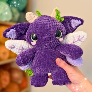 May include: A handmade, plush purple creature with large, expressive eyes and small, green leaf-like accents. It has wings with lavender and white details, and small beige horns. The toy is soft and cuddly, perfect for a child's room or as a unique gift.