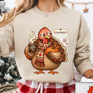 May include: A beige crewneck sweatshirt featuring a graphic of a turkey wearing a maple leaf patterned scarf, holding a mug labeled NOPE, and standing next to a November calendar with the 26th circled, paired with red and white plaid pants.