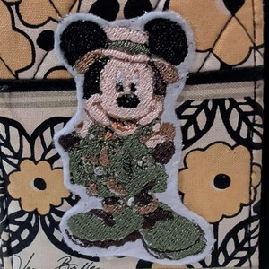 May include: A Vera Bradley wallet featuring a Mickey Mouse patch in a safari outfit. The wallet has a floral pattern in yellow and black, with a black zipper and the Vera Bradley signature.