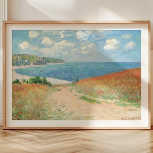 Op de afbeelding: Ingelijste kunstafdruk met een kustlandschap met een pad naar de zee. Het kunstwerk toont een heldere lucht met wolken, een blauwe oceaan en velden met rode bloemen. De lijst heeft een lichte houtkleur.