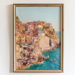 May include: Framed art print of a coastal Italian village. The artwork shows colourful buildings on a cliff overlooking the turquoise sea. The frame is gold, and the artwork is displayed on a white wall.