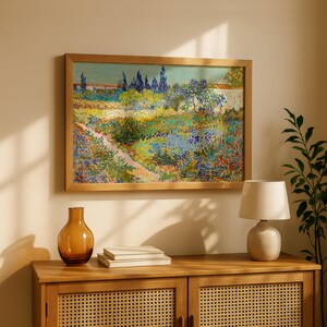 May include: A framed landscape painting with a wooden frame hangs above a wooden cabinet. The painting depicts a colorful garden scene with a path, trees, and flowers. A brown glass vase, a stack of books, and a small lamp sit on the cabinet.