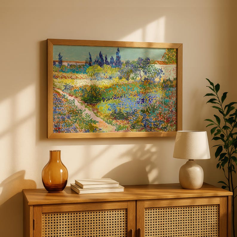 May include: A framed landscape painting with a wooden frame hangs above a wooden cabinet. The painting depicts a colorful garden scene with a path, trees, and flowers. A brown glass vase, a stack of books, and a small lamp sit on the cabinet.