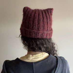 May include: A burgundy knitted beanie hat with cat ears. The hat has a ribbed design and a folded brim. The person is wearing a yellow t-shirt and a dark blue hooded sweatshirt.