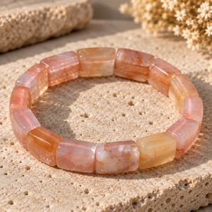 May include: A stretch bracelet composed of rectangular, polished beads in varying shades of translucent orange, peach, and pale pink. The beads feature natural internal inclusions and color variations, creating a smooth, glossy surface texture.