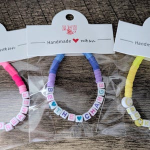 May include: Three handmade bracelets with colourful beads and positive messages. One bracelet says "DAY DREAMER" with a smiley face, another says "BE THE CHANGE" with heart beads, and the last says "BE KIND" with a smiley face.