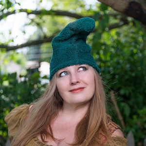 May include: A woman wearing a green fuzzy hat and a brown and red patterned top with a lace-up front. She is looking up and smiling.