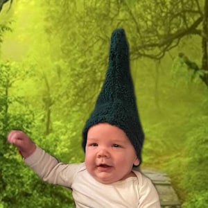 May include: A baby wearing a green gnome hat sits on a wooden path in a green forest setting.