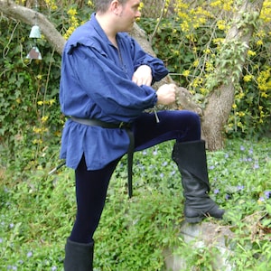 May include: A man wearing a blue shirt and dark blue trousers stands in a woodland setting. He is wearing black boots and a belt. The background is a lush green woodland with yellow flowers.