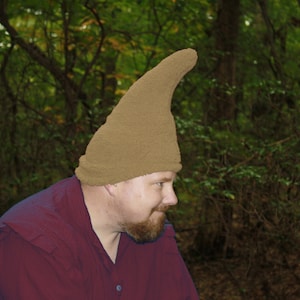 Może przedstawiać: Osoba w brązowym, puszystym kapeluszu z długim, spiczastym czubkiem. Kapelusz jest wykonany z miękkiego, pluszowego materiału. Osoba ma na sobie ciemnoczerwoną koszulę.