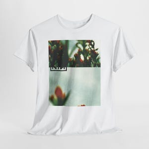 May include: White t-shirt with a blurred image of red and green plants. The image is split into two sections, with the word "NINE" in a black rectangle at the bottom of the top section. The shirt is made of a soft material.