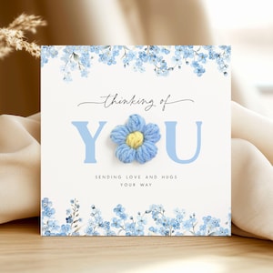 May include: A square greeting card featuring a blue knitted flower and the words "thinking of you." The card has a white background with blue floral accents and the text "sending love and hugs your way."