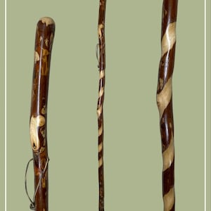 May include: A tall wooden walking stick featuring a polished finish with a distinct spiral pattern of dark brown and light tan wood grain. The staff has a smooth, rounded top and a small leather cord loop attached near the upper section of the shaft.