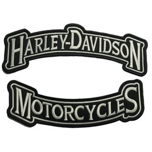 Könnte beinhalten: Zwei schwarz-weiße Harley-Davidson-Aufnäher. Der obere Aufnäher trägt in weißer, gewölbter Schrift die Aufschrift "HARLEY-DAVIDSON". Der untere Aufnäher trägt in weißer, gewölbter Schrift die Aufschrift "MOTORCYCLES". Beide Aufnäher haben einen schwarzen Rand.