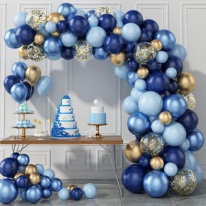 Puede incluir: Una escena festiva con un arco de globos en tonos azules y dorados. Una mesa está adornada con una tarta de varios pisos y otros postres. Globos y decoraciones adicionales completan el ambiente festivo.