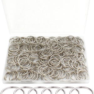 Puede incluir: Un recipiente de plástico transparente lleno de numerosos llaveros de color plateado. Los llaveros son redondos y de metal. Algunos llaveros son visibles en la parte inferior de la imagen, fuera del recipiente.